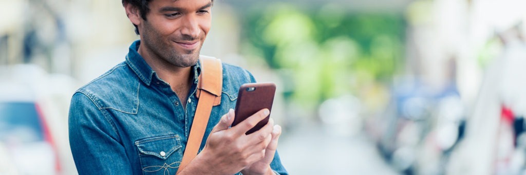A man in a denim shirt smiles down at his mobile phone.